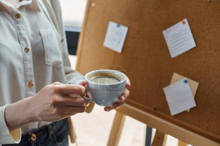 Une femme d'affaires dans un espace de bureau moderne, savourant une tasse de café près de son espace de travail.