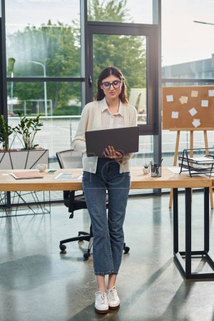 Une femme d'affaires se tient confiante dans un bureau moderne, travaillant sur son ordinateur portable près de son espace de travail alors qu'elle incarne le concept de franchise.