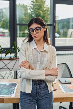 Une femme élégante avec des lunettes se tient en confiance près d'une table dans un cadre de bureau moderne, incarnant l'essence d'un concept d'entreprise de franchise.