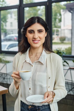A businesswoman in a modern office setting gracefully holds a cup of coffee and a plate, taking a break from her work.