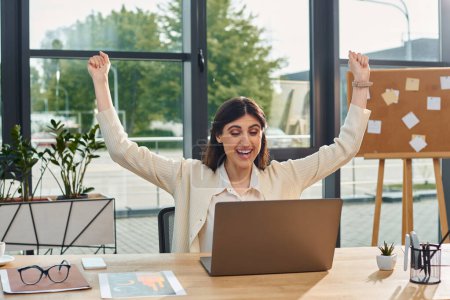Una mujer de negocios sentada en un escritorio con los brazos levantados para celebrar un concepto de franquicia exitoso en un entorno de oficina moderno.
