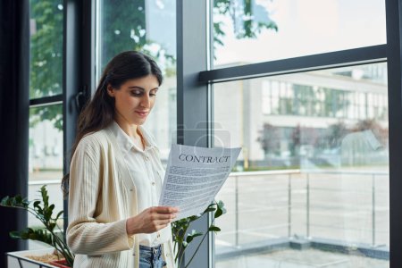Eine moderne Geschäftsfrau liest einen Vertrag, während sie an einem Fenster in ihrem Büro steht, umgeben von einem Franchise-Arbeitsplatz.