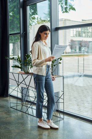 A businesswoman stands by a window, holding a contract in a modern office space, deep in thought.