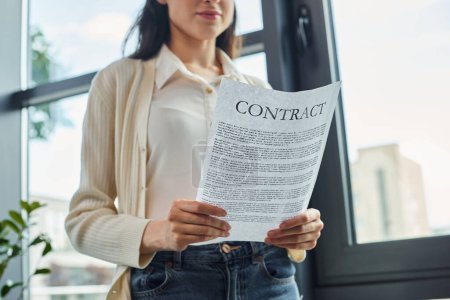 A businesswoman stands by a window, holding a contract in a modern office setting.