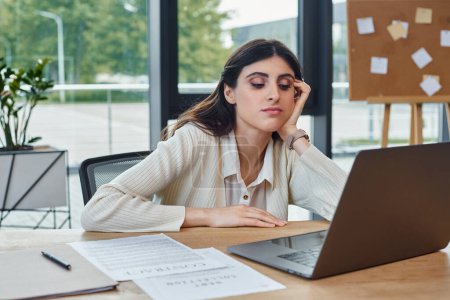 Una mujer de negocios se sienta en una mesa en una oficina moderna, enfocada en su computadora portátil, encarnando el concepto de una franquicia próspera.