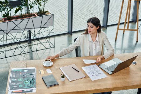 Foto de Una mujer de negocios se sienta en su escritorio rodeada de una computadora portátil y papeles, centrada en su trabajo de franquicia en una oficina moderna. - Imagen libre de derechos