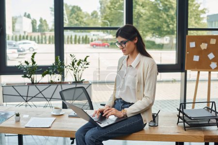 Téléchargez les photos : Une femme d'affaires assise sur une table à l'aide d'un ordinateur portable dans un bureau moderne, se concentrant sur le travail de franchise. - en image libre de droit