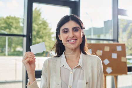 A modern businesswoman holds up a business card in front of a window in a sleek office space.
