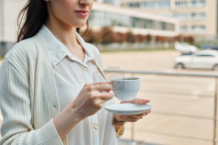 Una mujer de negocios sostiene con gracia una taza y un platillo al aire libre, encarnando un momento de calma en medio del bullicioso concepto de franquicia de una oficina moderna.