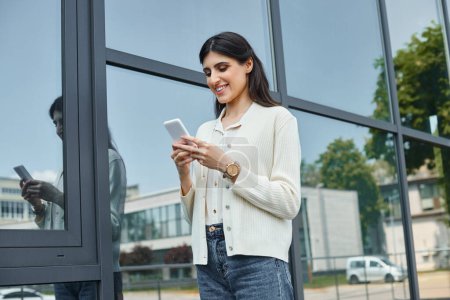 Une femme d'affaires concentrée se tient devant un bâtiment, naviguant sur son téléphone avec une expression réfléchie.