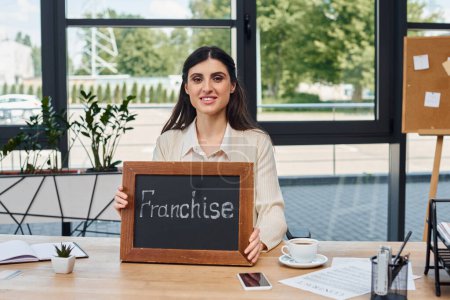 A modern businesswoman sits confidently at a table, holding a sign with a purposeful expression.