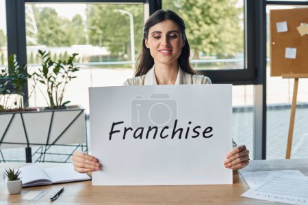 Eine Geschäftsfrau in einem modernen Büro sitzt an einem Tisch und hält ein Schild mit Franchise-Buchstaben in der Hand