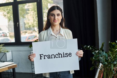 Una empresaria de oficina moderna sostiene con confianza un cartel que lee la franquicia en un gesto simbólico de emprendimiento.