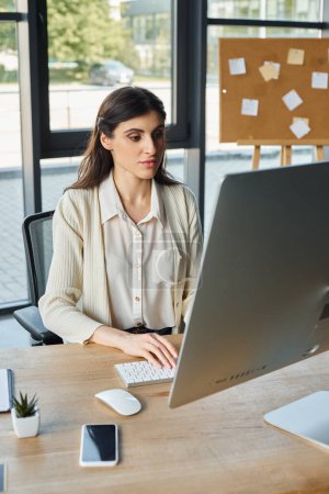 A determined businesswoman sits at her desk in a modern office, fully engaged with her computer.