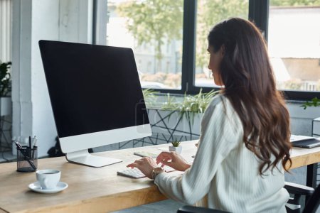 Eine Geschäftsfrau, mitten in der Arbeit, sitzt an ihrem Schreibtisch mit einem Computerbildschirm, der ihr Gesicht in einem modernen Büroumfeld erhellt..