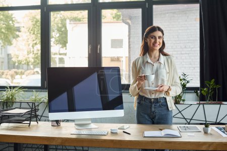 Eine moderne Geschäftsfrau steht an ihrem Computer und jongliert mit Aufgaben, während sie eine Tasse Kaffee in der Hand hält.