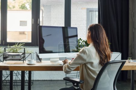 Una mujer de negocios inmersa en su trabajo, sentada en su escritorio en una oficina moderna, enfocada en la pantalla de la computadora.
