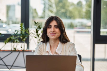 Una mujer de negocios se enfoca intensamente, sentada ante un portátil en una oficina moderna, encarnando determinación y compromiso.