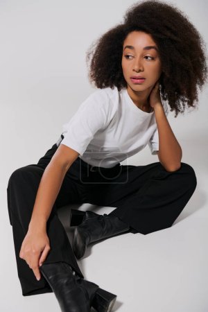 elegante mujer afroamericana en traje elegante sentado en el suelo y mirando hacia otro lado, concepto de moda