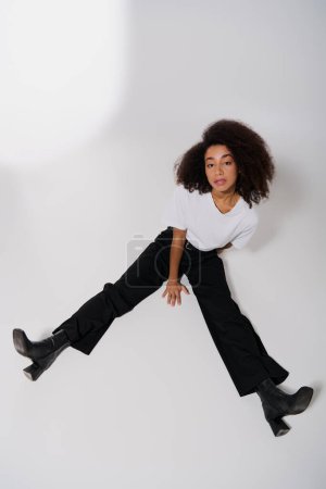 atractivo afroamericano modelo femenino sentado en el suelo y mirando a la cámara, concepto de moda