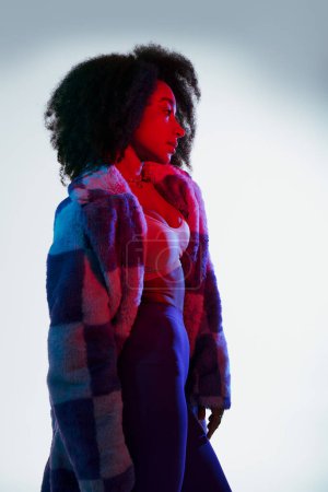 verführerische afrikanisch-amerikanische Frau in Kunstpelz, die in rotem und blauem Licht wegschaut, Modekonzept