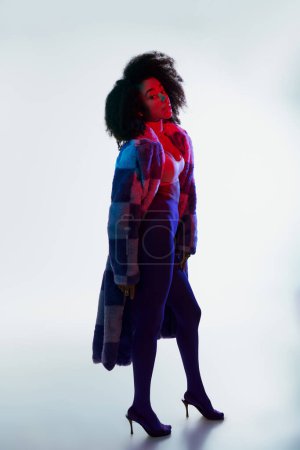 debonair afrikanisch-amerikanische Frau in Kunstpelz mit lockigem Haar, die in rotem und blauem Licht in die Kamera schaut
