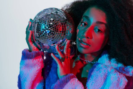 appealing stylish african american woman in faux fur posing with disco ball and looking at camera