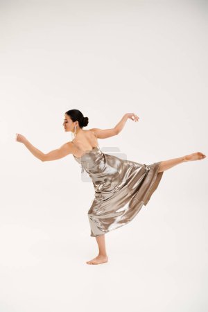 A young woman in a silver dress gracefully dances in a studio setting against a white background.