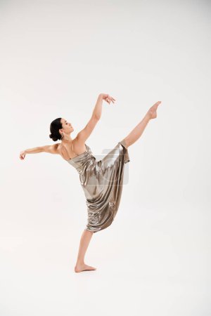 A graceful young woman in a long and shiny silver dress elegantly dances in a studio setting against a white background.