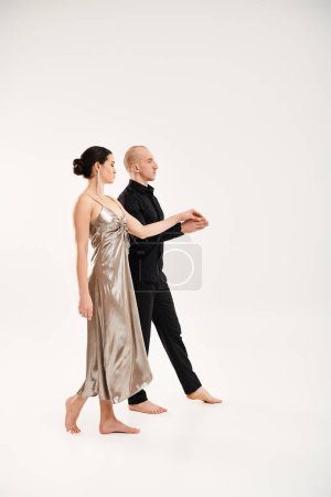 A young man in black and a young woman in a silver dress dance together in a studio shot on a white background.