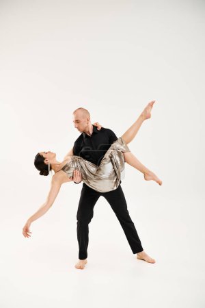 A young man in black and a young woman in a silver shiny dress dance together, performing acrobatic elements in a studio setting against a white background.