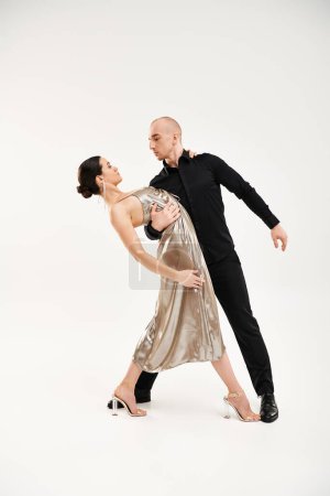 A young man in black and a young woman in a shiny dress perform acrobatic dance moves together in a studio setting against a white background.