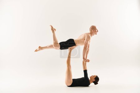 A shirtless young man performs a handstand atop another woman, both engaged in an acrobatic feat in a studio setting.
