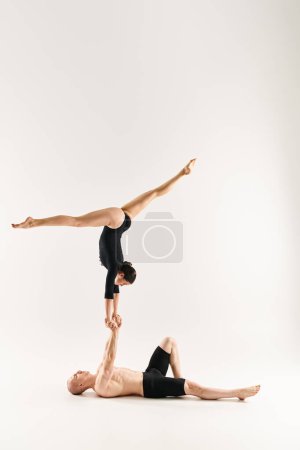 A shirtless young man and a young woman gracefully performing a handstand together in a studio setting against a white background.