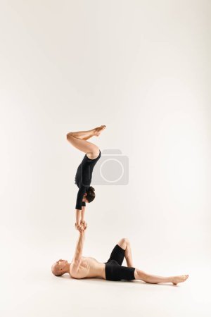 A young shirtless man and a woman perform a handstand, showcasing their acrobatic skills in a studio setting on a white background.