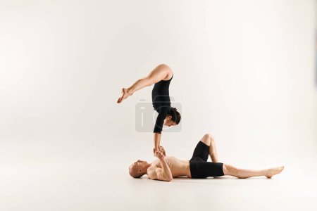 Shirtless man balances in a handstand on another man, showcasing strength and skill in acrobatics, white studio background.
