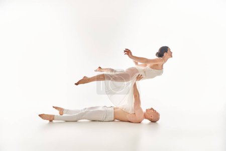 A shirtless young man and a woman in a white dress display grace and strength as they perform a split dance routine in a studio setting.