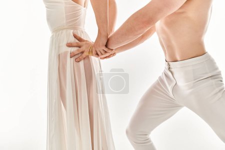 Un hombre joven, sin camisa y una mujer joven con un vestido blanco de pie entrelazado, tomados de la mano con gracia en una pose de baile.