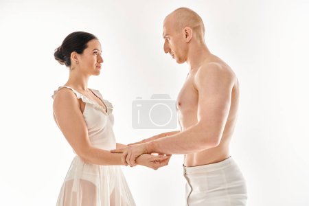 A shirtless young man and a woman in a white dress perform dance moves together in a studio against a white background.
