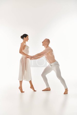 Ein hemdloser Mann und eine Frau im weißen Kleid führen gemeinsam in einem Studio-Setting akrobatische Tanzbewegungen aus.