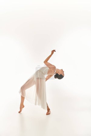 A graceful young woman in a flowing white dress performs in a studio setting against a white background.