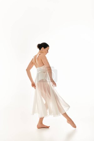 Una mujer joven en un vestido blanco que fluye con gracia baila en un entorno de estudio sobre un fondo blanco.
