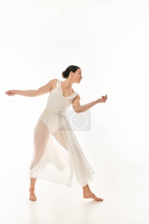 Une jeune femme respire l'élégance et la grâce en dansant dans une robe blanche fluide dans un décor de studio sur un fond blanc.