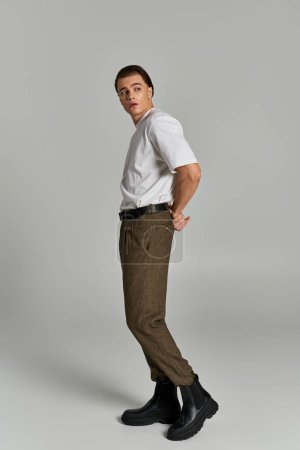 stylish young man in t shirt and brown pants posing attractively on gray backdrop and looking away