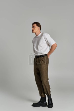 stylish young man in t shirt and brown pants posing attractively on gray backdrop and looking away