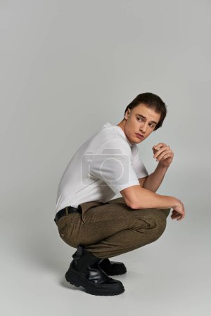handsome elegant male model in fashionable attire squatting and looking away on gray backdrop