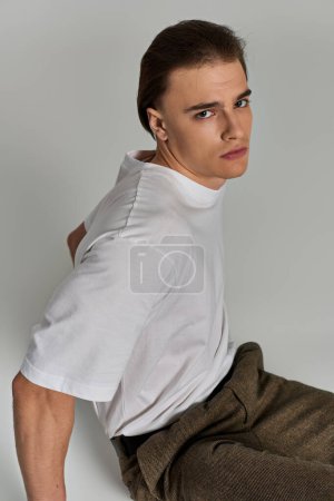 appealing polished young man in stylish clothing sitting on gray background and looking at camera