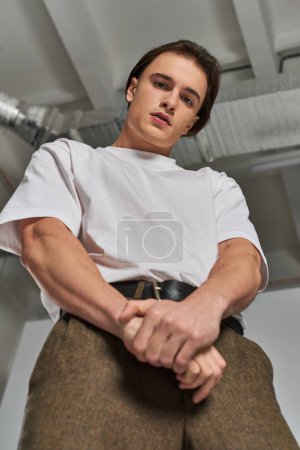 Foto de Modelo masculino joven de moda en pantalones sofisticados mirando a la cámara mientras está sobre fondo gris - Imagen libre de derechos