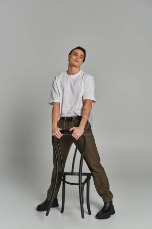 atractivo joven en traje de moda sentado atractivamente en la silla y mirando hacia otro lado en el fondo gris