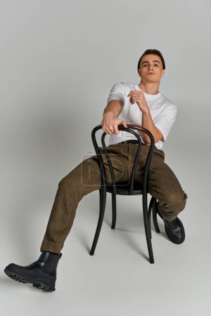 schöner stylischer Mann in Debonair-Kleidung sitzt auf einem Stuhl und schaut in die Kamera vor grauem Hintergrund